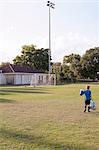 Rear view of boy football player carrying bag of footballs on practice pitch