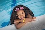 Portrait of girl with curly black hair hanging on edge of swimming pool, looking at camera smiling