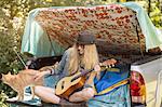 Young woman petting cat and playing ukulele whilst camping in pick up boot