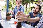 Young man and woman outdoors at a cafe, one trying to recork a bottle of wine.
