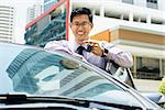 Young adult chinese businessman holds keys of new car, leaning on auto door. He smiles and looks proud at camera, showing the keys. Waist up portrait