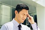 Portrait of asian businessman with eyesight problem, rubbing temple with pain expression. The man stands in a street against office buildings and holds eyeglasses