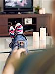 Young caucasian woman laying on sofa with colourful socks. She puts her feet on table and relaxes. The girl watches TV and holds remote control. Focus on socks