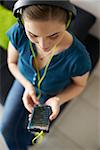 Beautiful woman relaxes on sofa and watches podcast on mobile phone, listening with green big earphones. High angle view