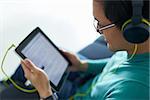 Chinese man relaxes on sofa and watches podcast on tablet pc, listening with green big earphones. Copy space