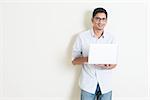 Portrait of handsome casual business Indian guy using laptop computer, standing on plain background with shadow, copy space at side.