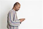 Side view of mature casual business Indian man using smartphone, mobile apps concept, standing on plain background with shadow.