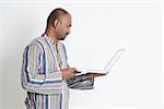 Side view of mature casual business Indian man using laptop computer, looking at pc screen, standing on plain background with shadow.