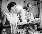 1950s SMILING HUSBAND IN APRON IN KITCHEN WASHING DISHES AS HAPPY WIFE DRIES