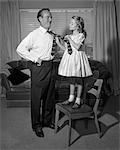 1950s CONFIDENT LITTLE GIRL STANDING ON CHAIR TYING NECKTIE FOR HER SMILING FATHER