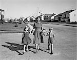 1950s SMILING HAPPY MOTHER HOLDING HANDS SON AND DAUGHTER WALKING ACROSS SUBURBAN HOUSING DEVELOPMENT STREET LOOKING AT CAMERA