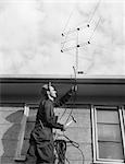1950s 1960s MAN WORKER ON LADDER RUNNING TELEVISION CABLE TO ANTENNA ON ROOF OF HOUSE