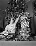 1950s SMILING YOUNG WOMAN SITTING UNDER CHRISTMAS TREE STACKING TALL PILE OF GIFT WRAPPED PRESENTS