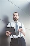 Stylish businessman using smartphone and digital tablet leaning against office wall