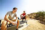 Two male friends, sitting on bench, on cliff top, looking at view