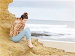 Young woman wrapped in shawl sitting on cliffside, Point Addis, Anglesea, Victoria, Australia
