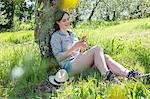 Young woman sitting leaning against tree using smartphone looking down smiling