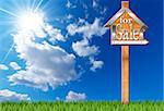 Wooden and metallic sign in the shape of house with text for sale and wooden pole. For sale real estate sign on blue sky with clouds, sun rays and green grass