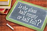 Is the glass half empty or half full question on a slate blackboard with a white chalk and a stack of books against rustic wooden table