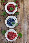 fresh raspberry, red currant and blueberry with green leaves in cups on wooden table, top view