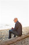 Bald man sitting on a log on a sandy beach.