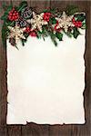 Christmas background border with gold snowflake bauble decorations, holly, ivy, cedar cypress and fir on parchment paper over old oak wood.