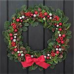 Christmas wreath with red bow and bauble decorations, holly, mistletoe and winter greenery over dark blue oak front door background.