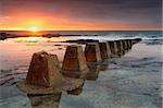 The sun rises at Coledale, casting its warm colours over the rockshelf with weathered and textured old pipeline holders in foreground.  There is a ship on the horizon.