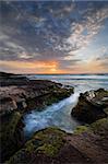 Beautiful coastal sunrise with tidal ocean flows into craggy eroded rock channel growing green and red moss or algae