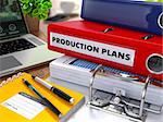 Red Ring Binder with Inscription Production Plans on Background of Working Table with Office Supplies, Laptop, Reports. Toned Illustration. Business Concept on Blurred Background.