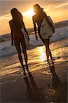 Rear view of two beautiful sexy young woman surfer girls in bikinis with white surfboards on a beach at sunset or sunrise