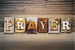 The word "PRAYER" written in rusty metal letterpress type sitting on a wooden ledge background.