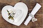 Romantic table place setting with white heart shaped porcelain bowls, antique silver fork with flower blossom and napkin with ring over old oak background.