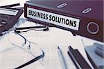 Ring Binder with inscription Business Solutions on Background of Working Table with Office Supplies, Glasses, Reports. Toned Illustration. Business Concept on Blurred Background.