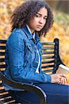 Sad thoughtful or depressed mixed race African American girl teenager female young woman drinking takeaway coffee outside sitting on a park bench in autumn or fall