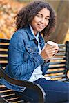 Beautiful happy mixed race African American girl teenager female young woman smiling and drinking takeaway coffee outside sitting on a park bench in autumn or fall