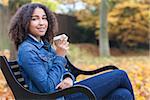 Beautiful happy mixed race African American girl teenager female young woman smiling and drinking takeaway coffee outside sitting on a park bench in autumn or fall