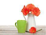 Orange gerbera flowers in pitcher and cup of drink on wooden table. Isolated on white background