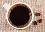 Top view of a white cup of black coffee. The cup stands on a linen fabric. There are three grains of coffee on the fabric near the cup. The fabric and the coffee beans are out of focus.