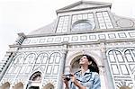 Low angle view of young woman using digital camera in front of church looking up, Piazza Santa Maria Novella, Florence, Tuscany, Italy