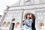 Low angle view of lesbian couple holding digital camera kissing on cheek in front of church, Piazza Santa Maria Novella, Florence, Tuscany, Italy