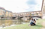 Lesbian couple sitting together on riverbank looking at digital camera in front of Ponte Vecchio and river Arno, Florence, Tuscany, Italy