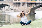 Young woman sitting on riverbank in front of Ponte Vecchio and Arno river using smartphone to take selfie, Florence, Tuscany, Italy