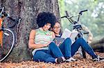 Mature woman and son sitting against park tree reading book and digital tablet
