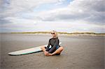 Senior woman relaxing on sand, surfboard beside her