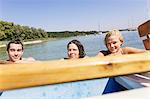 Friends with wet hair in lake holding onto boat looking at camera, Schondorf, Ammersee, Bavaria, Germany