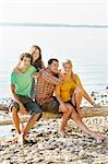 Friends sitting on driftwood next to shoreline looking at camera smiling, Schondorf, Ammersee, Bavaria, Germany