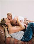 A woman lying on a sofa playing with a baby girl, kissing her cheek.