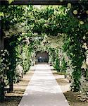 A garden path through an arch leading to an alcove with table and chairs, with climbing plants and white roses.