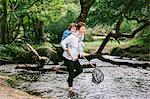 A woman giving a boy a piggyback and holding a shrimping net wading in a shallow stream.